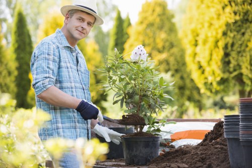 The image depicts a man working outdoors in a residential garden during daylight hours, surrounded by lush green trees with a bright and sunny weather. He is dressed in a light-colored straw hat, a blue checkered shirt, and gardening gloves, and is carefully handling a black plastic plant pot containing a young shrub or flowering plant. The garden features a well-maintained lawn area with dense, healthy grass in the foreground, and a prepared planting bed with dark, moist soil. In the background, there are several empty plant pots stacked neatly, along with gardening tools and supplies placed on a garden table or work surface. The scene emphasizes outdoor gardening activities, such as planting or potting, within a landscaped yard that likely benefits from professional gardening services to maintain its tidy and healthy appearance, consistent with the services offered by Lawn Mowing Elephant and Castle, serving local residents in the London SE1 postcode area.